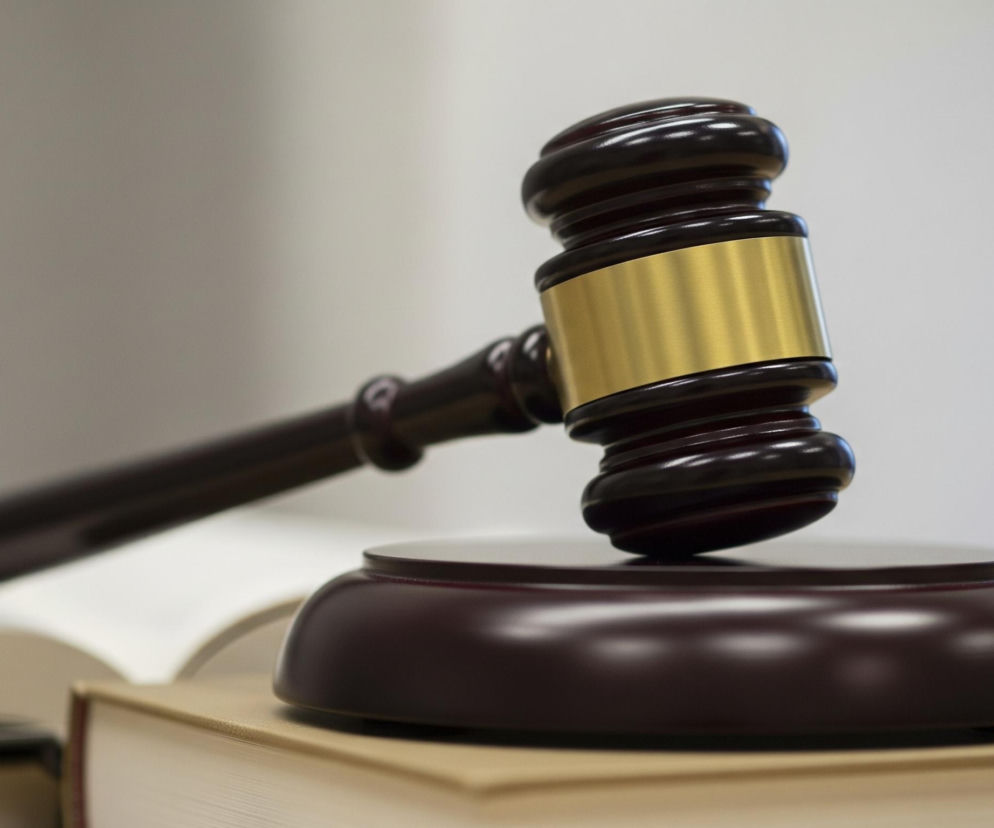 Młotek sędziowski wykonany z ciemnego, brązowego drewna, z połyskującą, złotą opaską wokół główki, spoczywa na owalnej podstawie w tym samym odcieniu drewna. Obiekt stoi na dwóch grubych książkach w jasnych, beżowych oprawach, z otwartą książką w tle po lewej stronie. Tło jest rozmyte i jasne, z widocznym cieniem po prawej stronie.