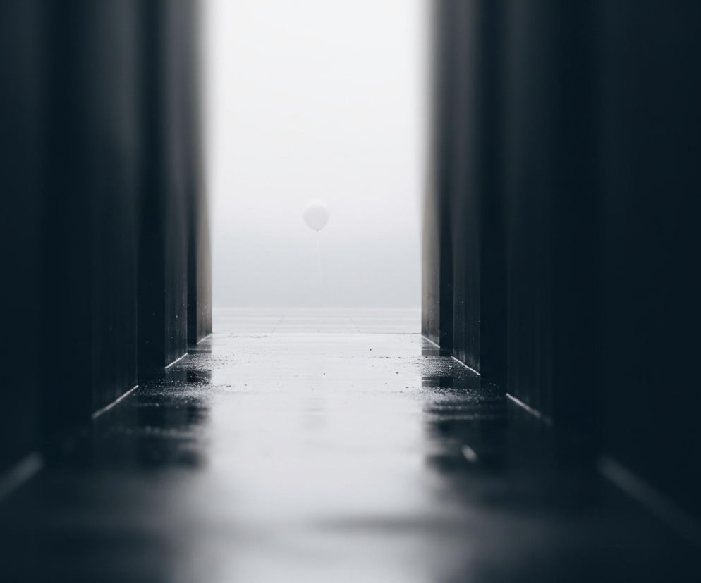 W centrum obrazu, w dalekim planie, unosi się biały, kulisty balonik, do którego przyczepiona jest cienka, biała nić zwisająca w dół. Balonik jest jedynym jasnym elementem w tle, które stopniowo rozjaśnia się od ciemnego do niemal białego, sugerując otwarta przestrzeń lub horyzont z zamglonym niebem lub wodą. Scena jest kadrowana przez dwa ciemne, pionowe bloki, które tworzą rodzaj korytarza lub przejścia, zbiegając się perspektywicznie ku środkowi. Podłoga korytarza jest ciemna i błyszcząca, z widocznymi refleksami światła, co wskazuje na mokrą powierzchnię, prawdopodobnie po deszczu.