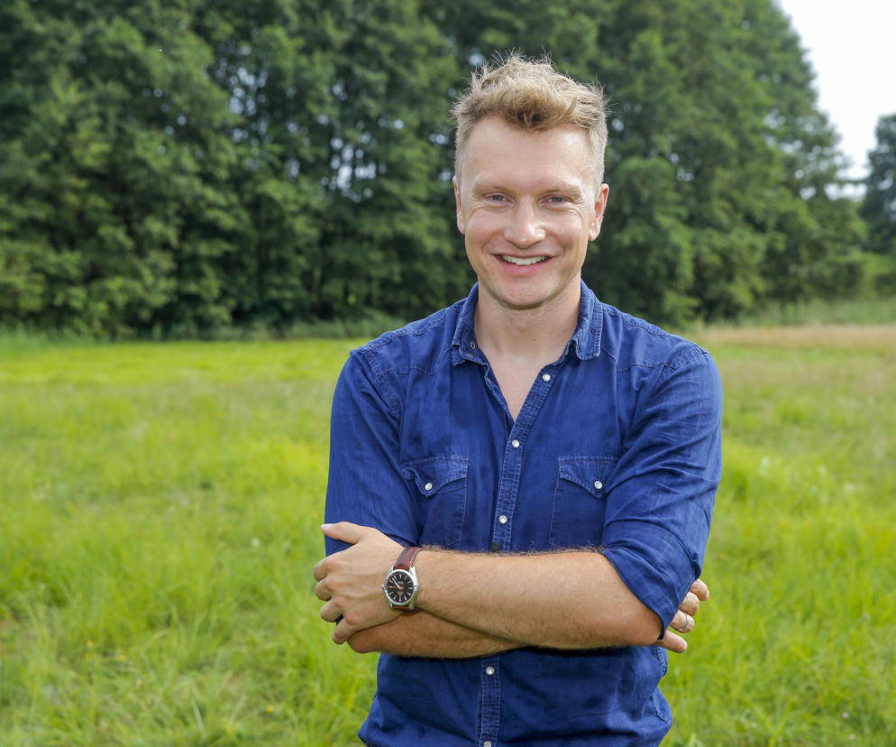 Portret uśmiechniętego Jakuba Wesołowskiego w denimowej koszuli, stojącego na zielonej łące z drzewami w tle, symbolizujący jego przemianę z bożyszcza nastolatek w dojrzałego mężczyznę. Więcej o metamorfozie aktora przeczytasz na naszym portalu.