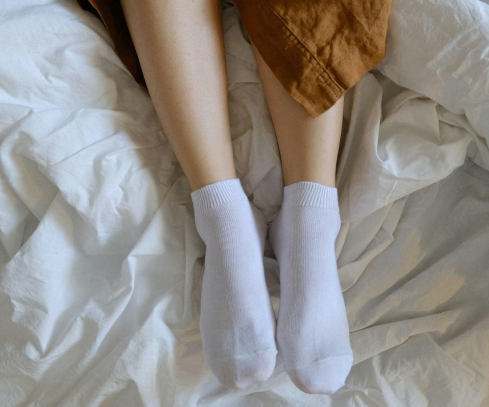 Damskie nogi w białych skarpetkach leżące na białym, pogniecionym prześcieradle, z brązowym materiałem na górze, symbolizujące ulgę dla zmęczonych nóg. Czytaj o naturalnym drenażu na Poradnik Zdrowie.