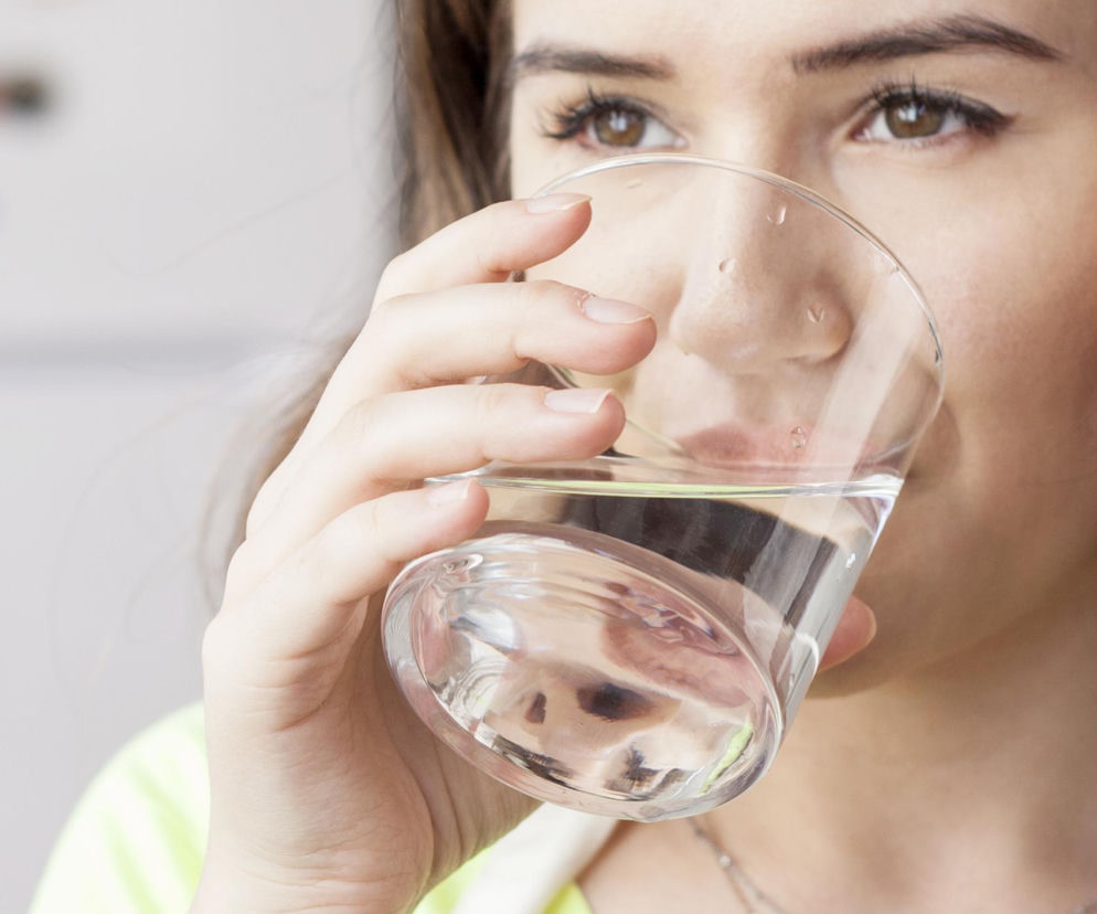 Kobieta pije wodę ze szklanki, nawadniając organizm. To symboliczne ujęcie w kontekście problemu suchości w ustach i kserostomii. Na Poradnik Zdrowie znajdziesz informacje o przyczynach i leczeniu tego stanu oraz domowych sposobach łagodzenia objawów.