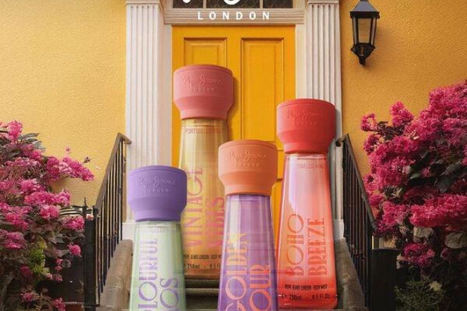 Cztery różnokolorowe mgiełki perfumowane Pepe Jeans Portobello Road ustawione na schodach żółtego budynku z kwitnącymi różowymi krzewami po bokach. Możesz przeczytać więcej o tej kolekcji na naszym portalu.