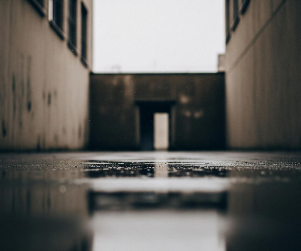 Ciemne, wilgotne przejście między budynkami, ujęte z bardzo niskiej perspektywy. Podłoże jest pokryte licznymi kroplami wody, które odbijają światło i tworzą lustrzane refleksy, szczególnie w dolnej części kadru. Po obu stronach widoczne są ściany budynków o matowej, beżowo-szarej barwie, z niewyraźnymi elementami architektonicznymi w górnej części. W oddali, na końcu przejścia, znajduje się otwór drzwiowy lub bramy, z którego wydobywa się jaśniejsze światło, rozmyte i nieostre. Całość utrzymana jest w chłodnej palecie barw, z dominującymi brązami, szarościami i ciemnym niebieskim odcieniem w odbiciach.