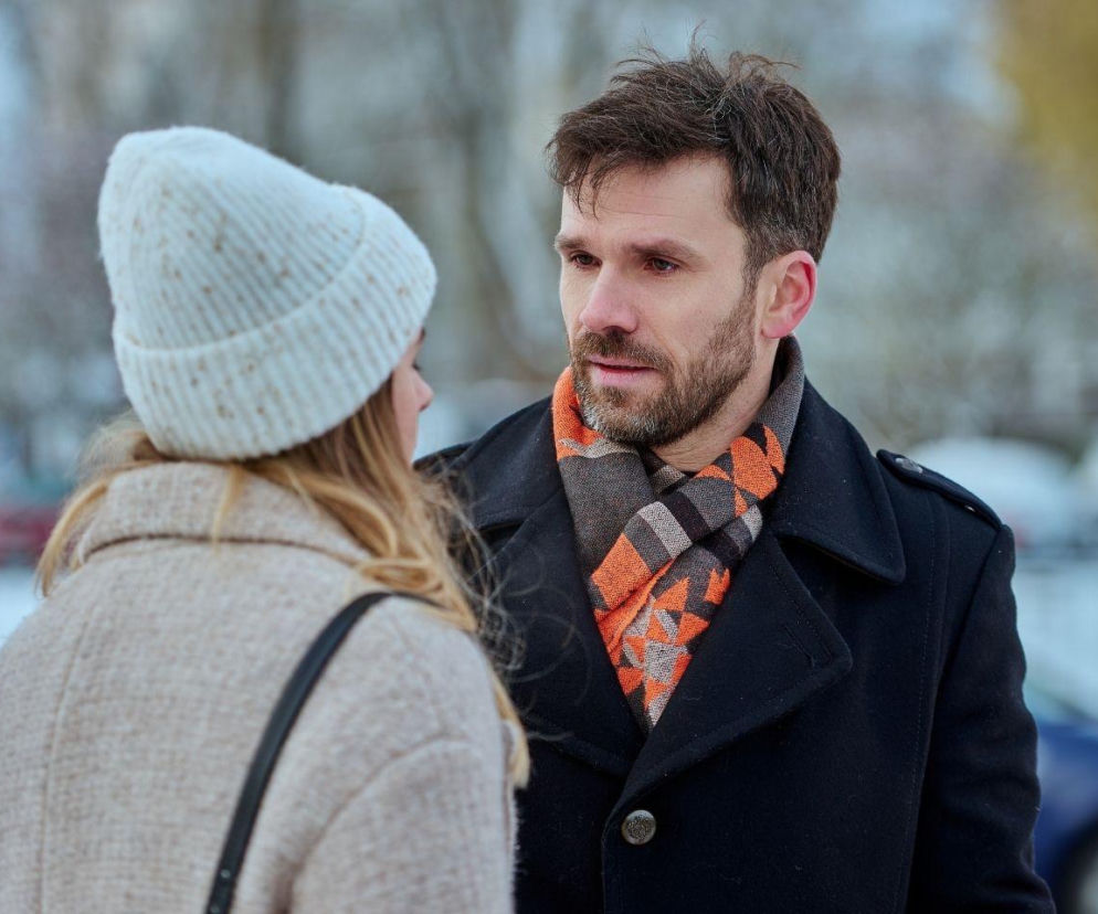 Mężczyzna z brodą w ciemnym płaszczu i wzorzystym szaliku patrzy na kobietę w jasnej czapce i płaszczu, uchwyconych w zimowej scenerii, co symbolizuje trudną rozmowę Elizy i Bartka z serialu Na Wspólnej. Więcej informacji o serialu Na Wspólnej znajdziesz na Super Seriale.