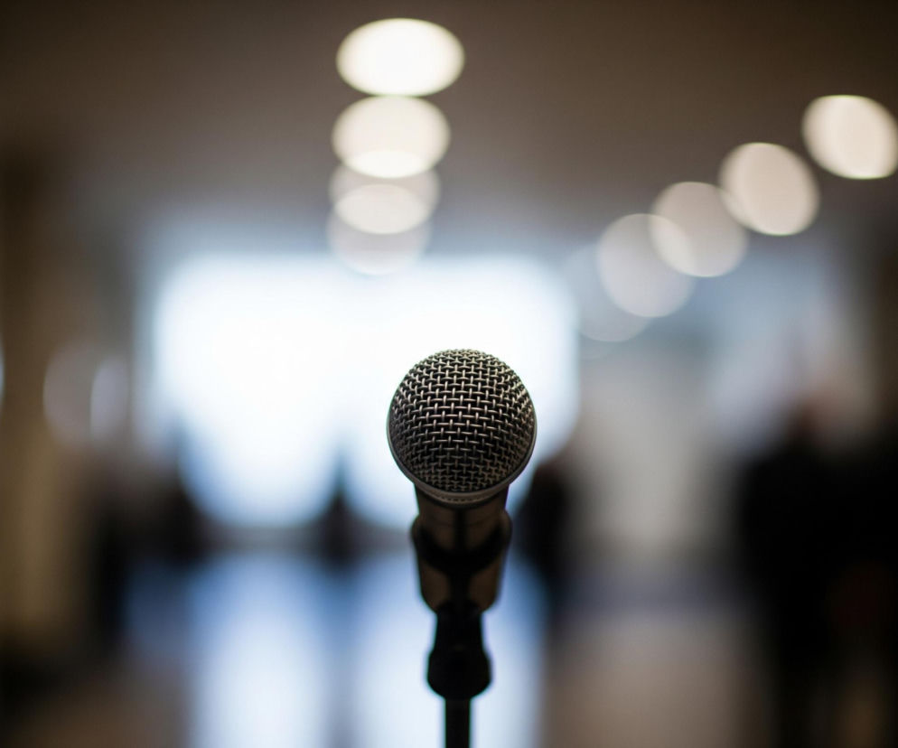 Na pierwszym planie, centralnie, stoi srebrno-czarny mikrofon na statywie, jego metalowa, siatkowa główka jest ostro zarysowana. Tło jest rozmyte, tworząc efekt bokeh, z niewyraźnymi, jasnymi punktami światła o różnych rozmiarach, rozmieszczonymi nieregularnie. W oddali widać zarysy postaci ludzkich oraz fragmenty wnętrza, prawdopodobnie korytarza lub dużej sali.