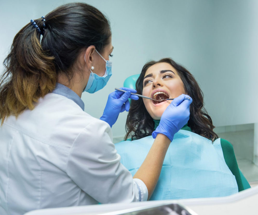 Dentystka w masce medycznej i niebieskich rękawiczkach bada zęby młodej pacjentki za pomocą lusterka stomatologicznego. Kobieta leży na fotelu dentystycznym z otwartymi ustami. Dowiedz się więcej o aktualnych cenach u dentysty na Poradnik Zdrowie.