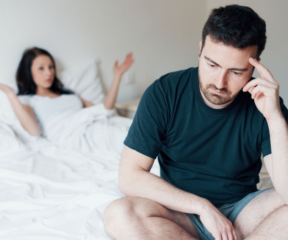 Zmartwiony mężczyzna siedzi na łóżku, trzymając dłoń na czole, podczas gdy w tle rozmazana sylwetka kobiety wyraża frustrację. Scena odzwierciedla problem dysfunkcji seksualnych, o których czytaj na portalu Poradnik Zdrowie.