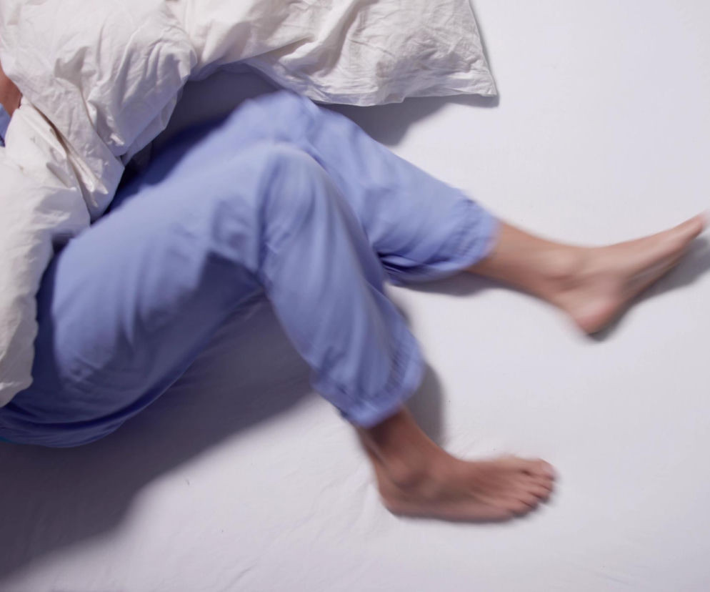 Osoba w niebieskiej piżamie, zakryta białą kołdrą, porusza niespokojnie nogami w łóżku na tle jasnej pościeli. Dynamiczne ujęcie podkreśla problem zespołu niespokojnych nóg, o którym więcej przeczytasz na portalu Poradnik Zdrowie.