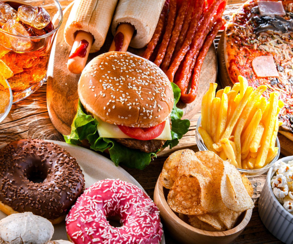 Stół zastawiony różnorodnymi produktami wysoko przetworzonymi: hamburgerem, hot dogami, pizzą, frytkami, chipsami, popcornem, pączkami i napojami gazowanymi, symbolizującymi niezdrową dietę. Odkryj na Poradnik Zdrowie, jak wpływa ona na mikrobiom jelitowy i samopoczucie.