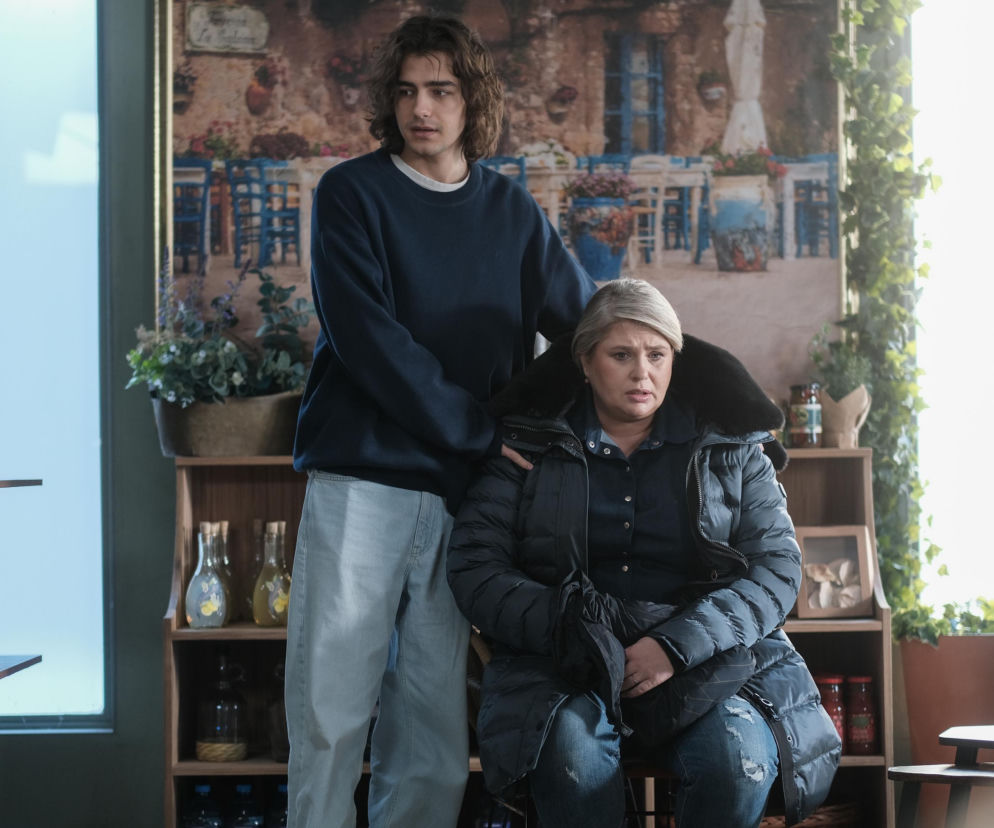 Kadr z Barw szczęścia przedstawiający Aldonę (Elżbieta Romanowska) i Aleksa (Hubert Wiatrowski) w sklepie Grzelaków. Aldona siedzi w puchowej kurtce z zmartwioną miną, a Aleks stoi za nią, trzymając rękę na jej ramieniu, wyrażając wsparcie. Oboje wyglądają na przejętych sytuacją po zatruciu klienta grzybami. Więcej o dramacie Grzelaków na Super Seriale.