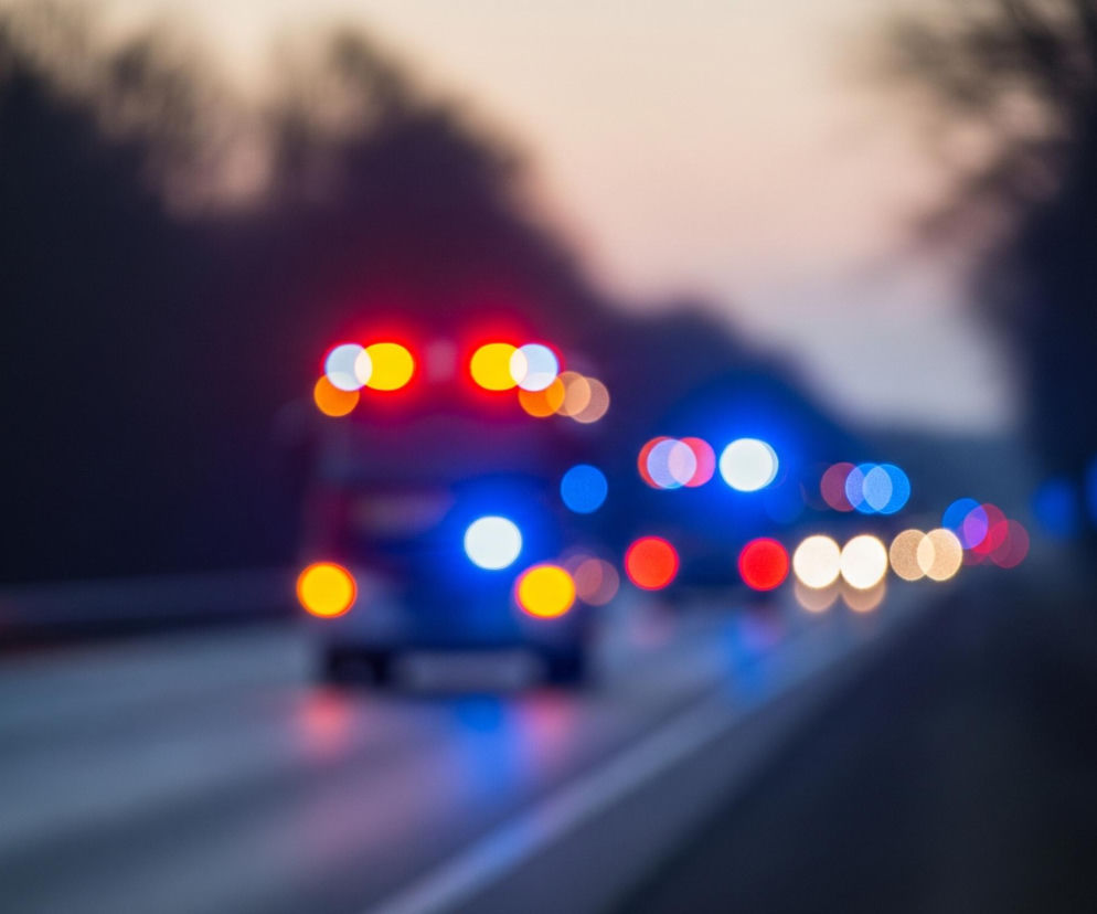 Rozmyte światła pojazdów poruszają się po dwupasmowej drodze w ciemnym otoczeniu. Głównym obiektem jest pojazd jadący w głąb kadru, z wyraźnie widocznymi, rozmazanymi czerwonymi i żółtymi światłami na dachu oraz niebieskimi i pomarańczowymi światłami z tyłu. Droga ma szarą nawierzchnię z jasnymi liniami oddzielającymi pasy ruchu, po prawej stronie widać rozmyty biało-czerwony słupek drogowy. Tło stanowią ciemne, rozmyte kształty drzew i drzewostanów, a nad nimi jaśniejsze, szaro-różowe niebo, sugerujące zmierzch lub świt.