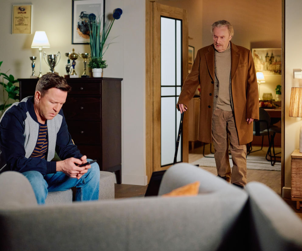 Wojtek siedzi na szarej kanapie w salonie, pochylony nad telefonem, zaniepokojony po groźbach Miśka, podczas gdy jego ojciec w jasnym płaszczu i z laską wchodzi do pokoju, by pomóc synowi. Artykuły o serialu Na Wspólnej dostępne na Super Seriale.