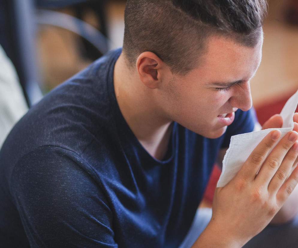 Młody mężczyzna w niebieskiej koszulce z krótkimi rękawami, trzymający chusteczkę higieniczną przy nosie, intensywnie kicha z zamkniętymi oczami. Ukazuje to powszechne zjawisko fosfenów po kichaniu, o którym więcej przeczytasz na Poradnik Zdrowie.