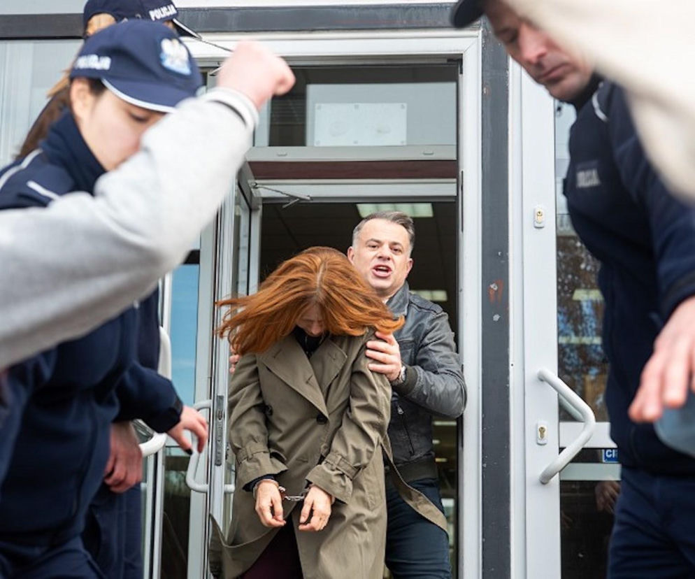 Kobieta z rudymi włosami w zielonym płaszczu i kajdankach na rękach, wyprowadzana z budynku przez mężczyznę w czarnej kurtce. W tle widać policjantów. To scena z serialu Pierwsza miłość odcinek 4141, gdzie Teresa trafia do aresztu. Więcej o losach bohaterów przeczytasz na portalu Super Seriale.