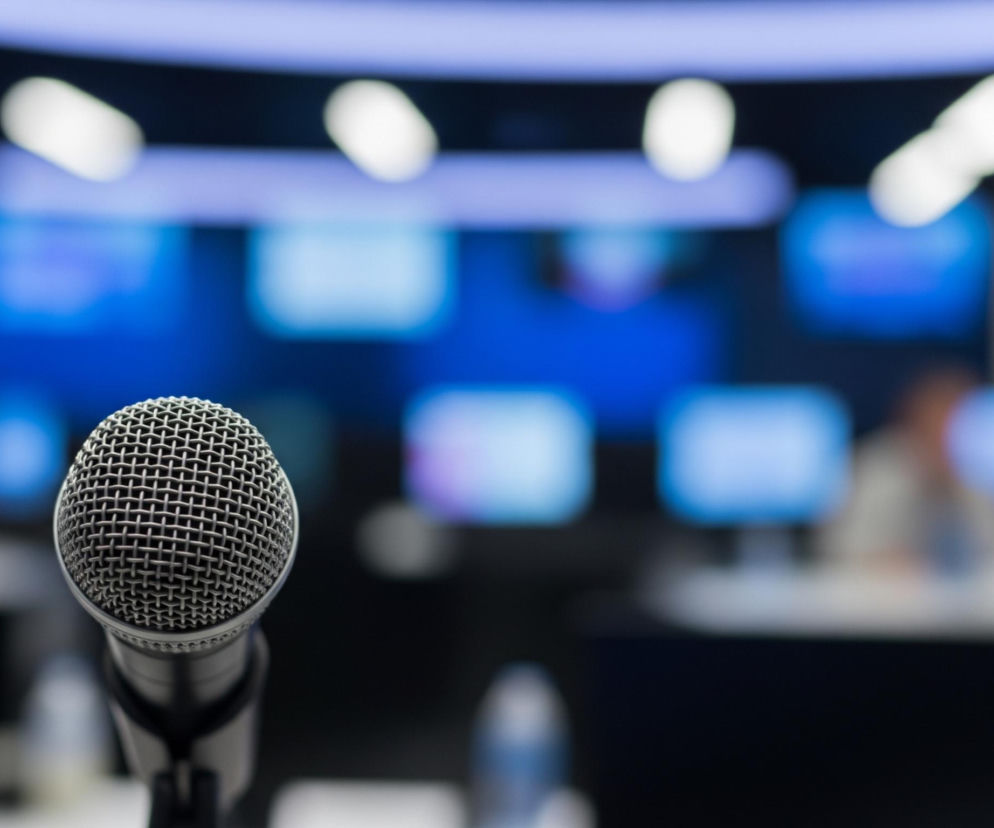 Mikrofon z metalową, siateczkową głowicą i czarnym, matowym korpusem znajduje się na pierwszym planie, lekko na lewo od centrum obrazu. Jest on ostry, kontrastując z rozmytym tłem, które przedstawia wnętrze pomieszczenia o niebiesko-fioletowej kolorystyce. W tle widoczne są niewyraźne, jasne prostokąty, przypominające ekrany komputerowe, oraz rozmyte postacie siedzące przy stole, co sugeruje salę konferencyjną lub studio. Górna część obrazu ukazuje jasne, podłużne źródła światła na suficie.