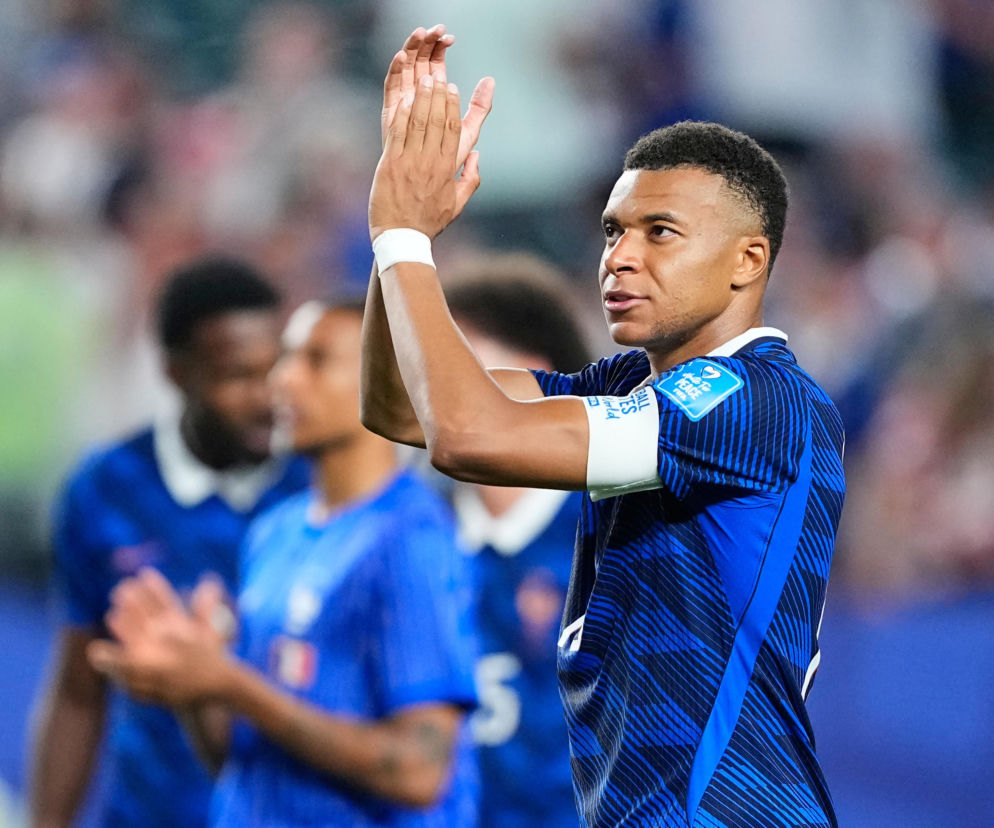 Kylian Mbappe z reprezentacji Francji w granatowej koszulce piłkarskiej klaska na boisku. W tle widać niewyraźne sylwetki innych zawodników. Więcej o meczu Francja – Irak na naszym portalu.