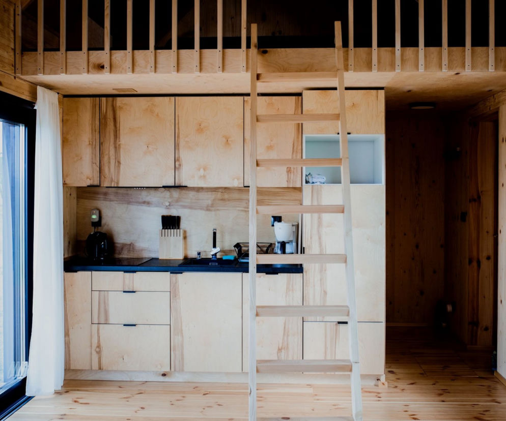 Wnętrze kompaktowego domu Hyttee z widocznym aneksem kuchennym z drewnianymi szafkami i czarnym blatem oraz drewnianą drabiną prowadzącą na antresolę. Po lewej duże okna tarasowe z białą zasłoną. Architektura Murator Plus to idealne miejsce, by dowiedzieć się więcej o tym innowacyjnym projekcie.