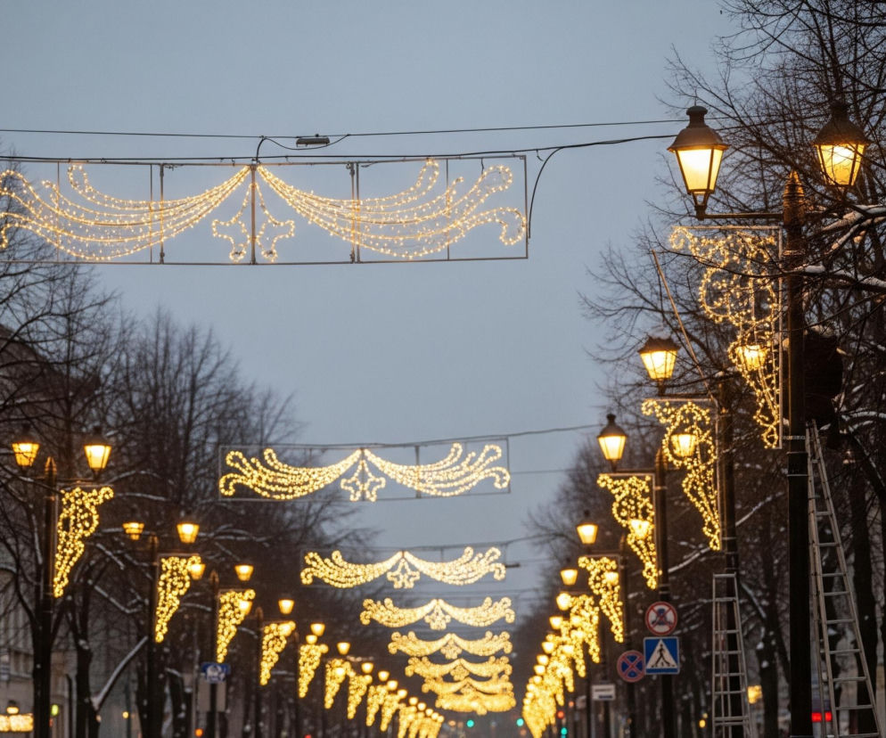 Ulica ozdobiona jest licznymi, ciepło żółtymi iluminacjami świetlnymi, rozwieszonymi nad jezdnią oraz na latarniach wzdłuż chodników. Na pierwszym planie, po prawej stronie, widać wysoką, aluminiową drabinę opartą o latarnię, a u jej podnóża stoją dwie osoby. Nagie drzewa po obu stronach ulicy pokryte są niewielką warstwą śniegu, podobnie jak część budynków widocznych w tle, a niebo jest pochmurne i szare.