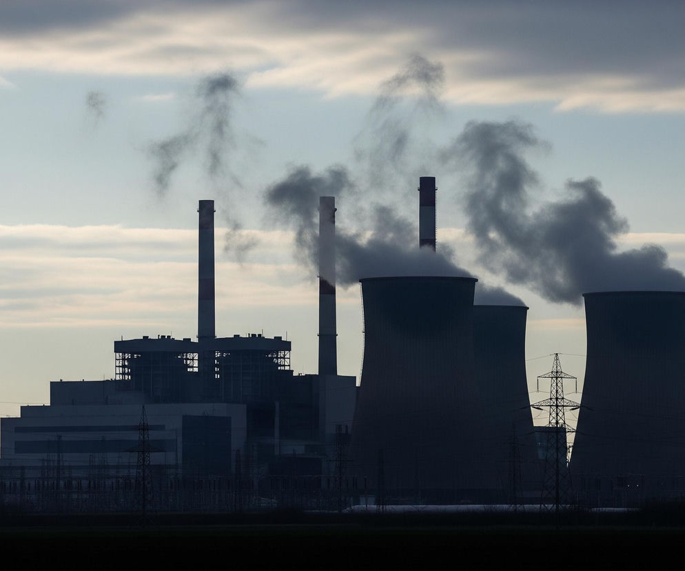 Centralnie usytuowana jest duża elektrownia w świetle zmierzchu, z dominującymi ciemnymi, szarymi i niebieskawymi odcieniami, tworzącymi sylwetkę. Widoczne są cztery kominy, z których trzy emitują gęsty, ciemny dym w różnych odcieniach szarości, rozprzestrzeniający się w niebo. Po prawej stronie znajdują się trzy ogromne chłodnie kominowe, dwie z nich również wydzielają obfite obłoki dymu. Na pierwszym planie, wzdłuż dolnej krawędzi obrazu, rozciąga się ciemne, płaskie pole, a nad nim liczne, smukłe wieże przesyłowe o ażurowej konstrukcji, niektóre z widocznymi przewodami, otaczają elektrownię, tworząc złożoną sieć. Niebo jest częściowo zachmurzone, z pasmami jasnych, szarawych chmur na tle bladoniebieskiego, niemal przezroczystego nieba.