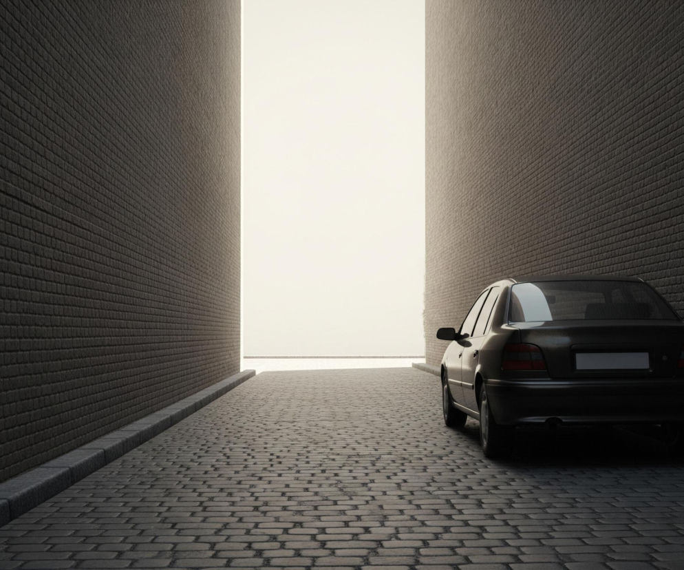 Ciemnoszary samochód osobowy jest zaparkowany na brukowanej ulicy, ustawiony prawym bokiem do widza. Ulica, wykonana z ciemnoszarych, nieregularnych kostek brukowych, prowadzi prosto w kierunku jasnego, białego światła na końcu wąskiego przejścia. Po obu stronach ulicy wznoszą się wysokie ściany zbudowane z cegieł w bardzo ciemnym odcieniu szarości, niemal czarnym, z lekkim, ciepłym refleksem od jasnego światła. Krawężniki oddzielają ściany od bruku.