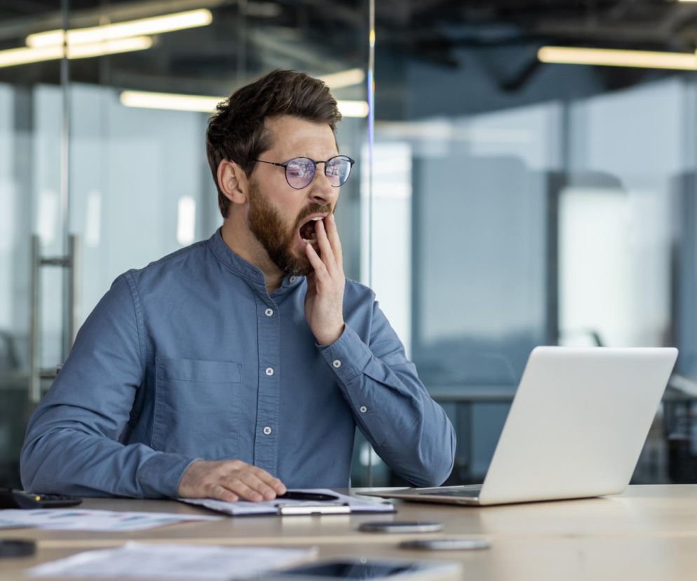Młody mężczyzna w okularach i błękitnej koszuli ziewa, zasłaniając usta dłonią, siedząc przed laptopem przy biurku w biurze. Męczące popołudniowe zmęczenie i senność to objawy spadku energii, o których przeczytasz na Poradnik Zdrowie.