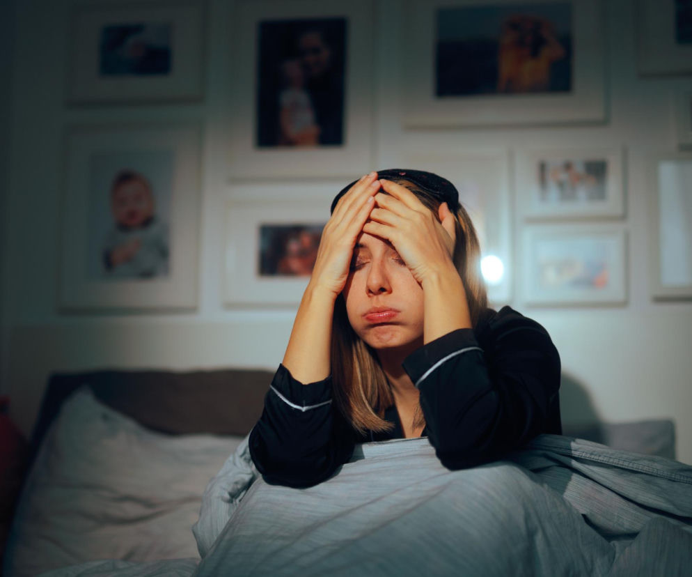 Zmęczona kobieta w piżamie i opasce na oczy siedzi w łóżku z rękami na głowie, symbolizując problemy ze snem przy cukrzycy. Tło sypialni z ramkami na zdjęcia podkreśla osobisty kontekst. O tym, jak poprawić sen przy cukrzycy, przeczytasz na Poradnik Zdrowie.