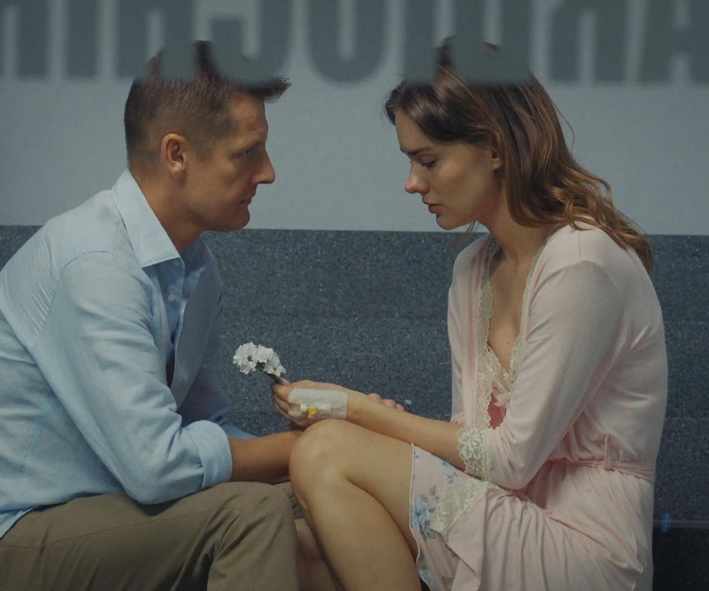Paweł (Rafał Mroczek) w jasnoniebieskiej koszuli i beżowych spodniach, siedzi naprzeciwko Franki (Dominika Kachlik) w różowym szlafroku z koronką, z wkłuciem wenflonowym na dłoni. W tle szare, kamienne schody. Paweł ofiarowuje Frankce białe kwiaty, oboje spoglądają na bukiecik, odzwierciedlając romantyczny, choć pełen obaw moment opisywany na portalu Super Seriale.