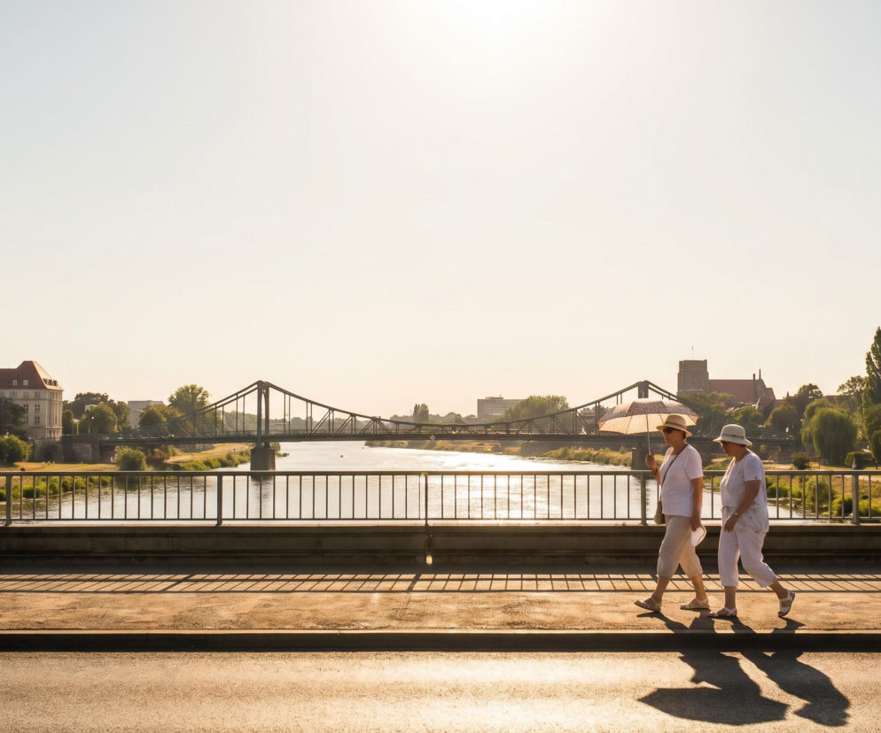 Dwie kobiety w jasnych ubraniach i kapeluszach idą po moście w słoneczny dzień, jedna z parasolką przeciwsłoneczną, co odzwierciedla falę upałów i rekordowych temperatur, o których przeczytasz więcej na naszym portalu. W tle widać rzekę i zabudowania.