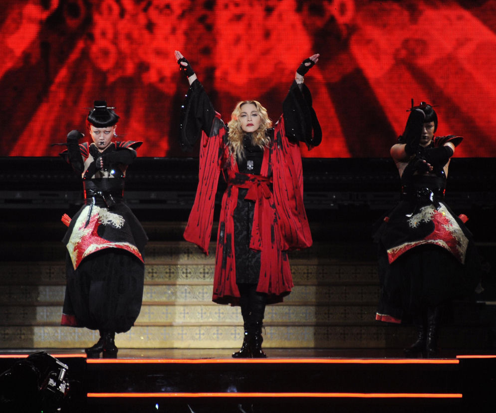 Madonna w czerwono-czarnym stroju z rozłożonymi ramionami, na scenie w otoczeniu dwóch tancerek w podobnych stylizacjach. Scena w tle rozświetlona czerwonym światłem. Przeczytaj o jej krytyce AI na naszym portalu.