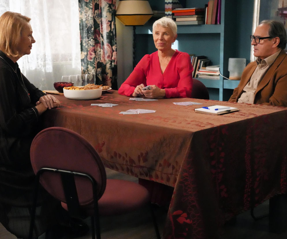 Krystyna, Wanda i Jerzy siedzą przy stole, grając w brydża. Wanda w czerwonej bluzce uśmiecha się, trzymając karty. Jerzy w brązowej marynarce i beżowej koszuli, obok niego Krystyna w czarnej sukience, obie panie skupione na grze. Na stole talie kart, miska z przekąskami i notatnik. Całość zdjęcia oddaje atmosferę towarzyskiego spotkania, o którym można przeczytać na portalu Super Seriale.