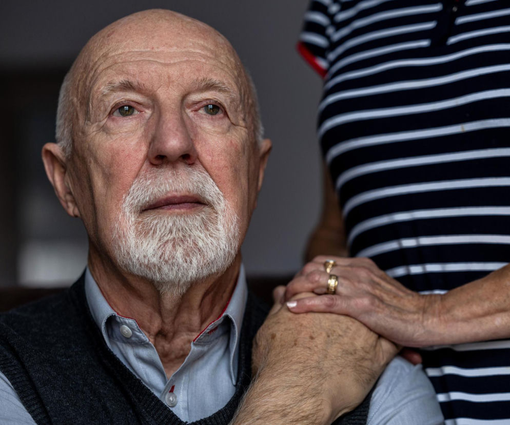 Starszy mężczyzna z białą brodą i wąsami, w szarej koszuli i swetrze, z zatroskanym wyrazem twarzy. Jego dłoń spoczywa na ręce kobiety, symbolizując wsparcie w chorobie Parkinsona. O pierwszych objawach i wsparciu przeczytasz na Poradnik Zdrowie.