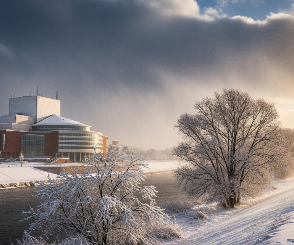 Duży budynek o ceglanych ścianach i przeszklonych, półokrągłych fasadach dominuje w lewej części kadru, jego biały, ośnieżony dach zlewa się z częściowo zasłoniętym niebem; poniżej płynie rzeka, po jej prawej stronie rozciąga się ośnieżony wał obsadzony drzewami, również pokrytymi grubą warstwą śniegu. Po prawej stronie wału widać ośnieżoną ścieżkę, którą podążają ludzie ubrani w zimowe kurtki, oraz rząd latarni. Niebo jest częściowo ciemnoszare, z widocznymi opadami śniegu, a częściowo rozświetlone złotym światłem zachodzącego słońca, tworząc kontrast między ponurym, zachmurzonym niebem po lewej stronie a jasnym, słonecznym po prawej.