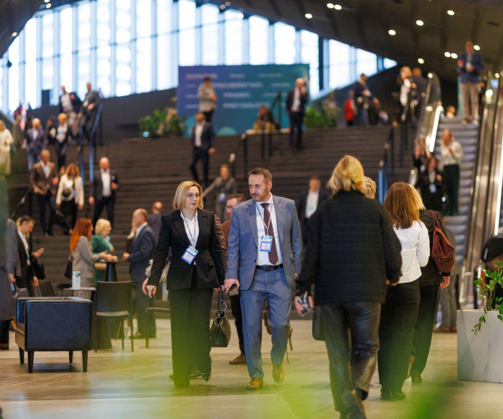 Uczestnicy XI Kongresu Wyzwań Zdrowotnych Health Challenges Congress w Międzynarodowym Centrum Kongresowym w Katowicach podczas przerw między sesjami. Osoby w strojach biznesowych, część z nich posiada identyfikatory. Na drugim planie widoczne schody i ruchome schody, a w tle duże okno. Poradnik Zdrowie informuje o rejestracji na to ważne wydarzenie.