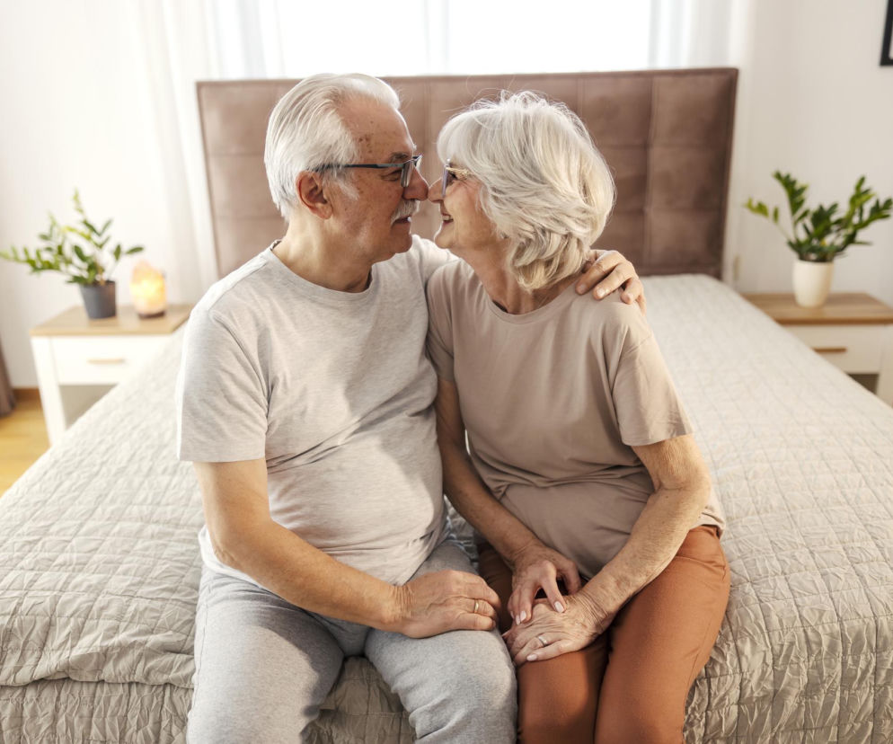 Starsze małżeństwo siedzące na łóżku w sypialni, z uśmiechem i czułością dotykające się nosami. Mężczyzna obejmuje kobietę ramieniem, a ich dłonie splatają się, co symbolizuje bliskość i intymność seniorów. Więcej o seksie po 70. roku życia na Poradnik Zdrowie.