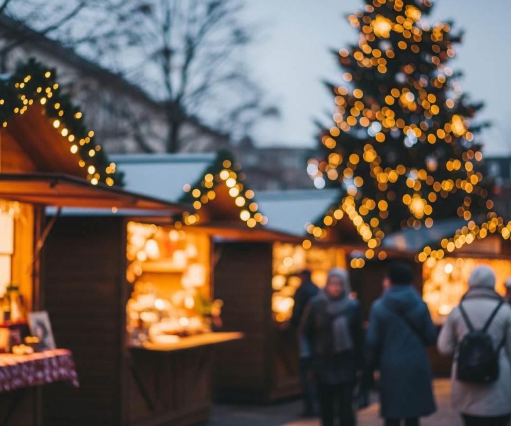 Scena przedstawia jarmark rozświetlony licznymi, ciepłymi, żółtymi światłami. W centralnej części widać duże, rozmazane drzewo ozdobione okrągłymi, jasnożółtymi lampkami. Po lewej stronie znajdują się drewniane stragany z dachami udekorowanymi girlandami świateł, przy czym jeden z nich, najbliżej widza, ma stół przykryty czerwonym obrusem z białym wzorem i półki z niewyraźnymi przedmiotami. W tle widoczne są sylwetki ludzi, spacerujących między straganami, oraz ciemne budynki i drzewa bez liści pod szarobłękitnym niebem, wszystko poza pierwszym planem jest rozmyte, co tworzy efekt bokeh.