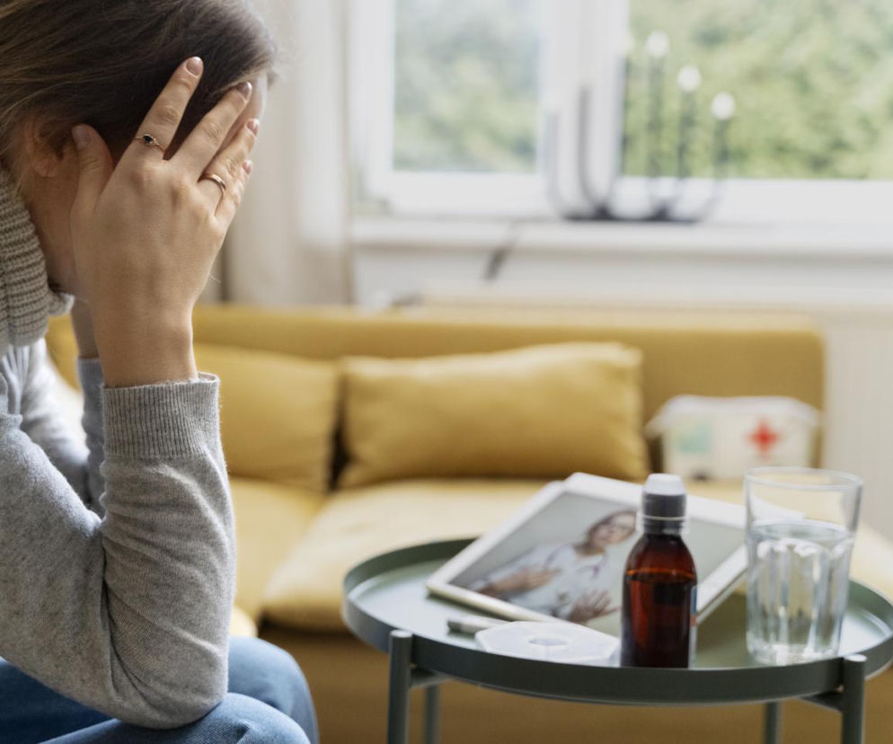 Kobieta z dłońmi na twarzy siedząca w szarym swetrze, obok na stoliku syrop i szklanka wody. Obraz symbolizuje zmartwienie związane z chorobą i zwolnieniem lekarskim. O zmianach w przepisach czytaj na Super Biznes.
