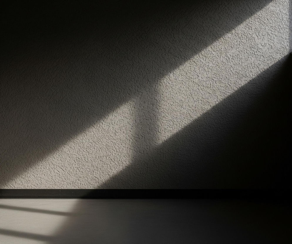 Wnętrze ciemnego, pustego pokoju o teksturowanych ścianach i gładkiej podłodze, oświetlone ukośnym pasem światła słonecznego padającego z prawego górnego rogu. Prawy górny róg ujawnia fragment okna z pionowymi szprosami i widokiem na jasne, rozmyte tło na zewnątrz. Na dole ścian widoczna jest ciemna, wąska listwa przypodłogowa, a światło słoneczne częściowo oświetla podłogę, tworząc kontrast między jasnymi i ciemnymi obszarami.