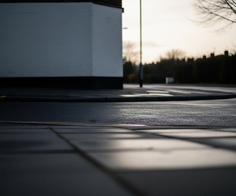 Chodnik z prostokątnych płyt rozciąga się na pierwszym planie, z jego ciemnymi powierzchniami odbijającymi jasne światło. Droga, lekko mokra i o ciemnoszarej nawierzchni, zakręca w prawo, z podwójną żółtą linią wyznaczającą krawędź. Po lewej stronie wznosi się biały budynek z ciemnym cokołem, a obok niego widoczna jest szara latarnia uliczna. W tle domy i bezlistne drzewa rysują się na jasnym, nieco rozmytym tle nieba.