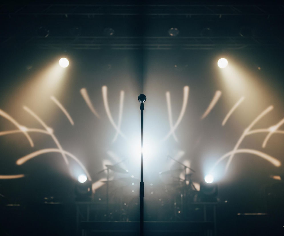 Centralnie, na pierwszym planie, stoi czarny mikrofon na statywie, intensywnie oświetlony od tyłu przez jasny reflektor, tworząc długą, białą smugę światła biegnącą w górę. Po obu stronach mikrofonu, w tle, widoczne są dwa okrągłe, ciepłe, żółto-białe reflektory emitujące szerokie stożki światła. Na ciemnej ścianie za mikrofonem widoczne są abstrakcyjne, białe wzory świetlne składające się z zakrzywionych linii, przypominających fragmenty okręgów. W tle, po lewej i prawej stronie, można dostrzec niewyraźne zarysy instrumentów muzycznych, prawdopodobnie perkusji i wzmacniaczy, częściowo zasłoniętych przez intensywne światło i dym.