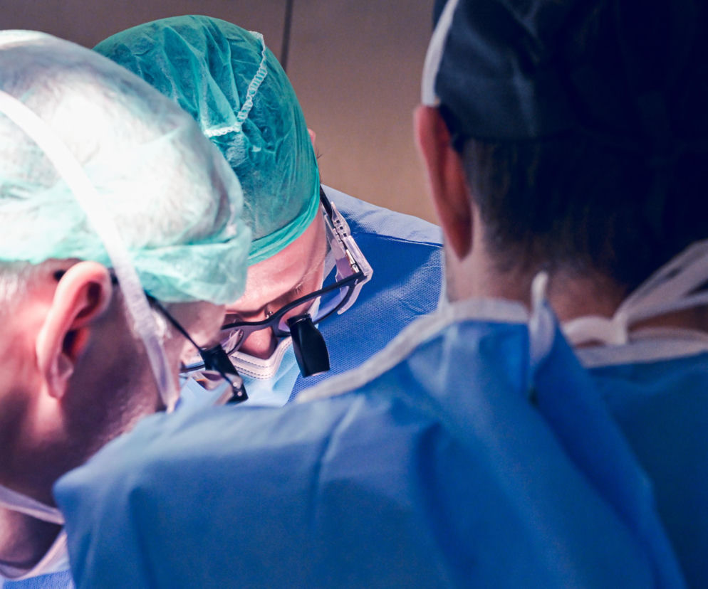 Trzech chirurgów w zielonych czepkach operacyjnych i niebieskich fartuchach, jeden z nich w okularach z lupą, pochylonych nad pacjentem podczas zabiegu. Temat zarobków lekarzy i chaosu w ochronie zdrowia omawiany jest na portalu Super Biznes.