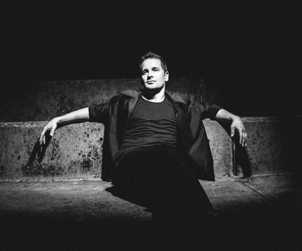 Krzysztof Zalewski siedzący na betonowym murku w czarno-białym kadrze, z otwartą marynarką i swobodną postawą, patrzy w dal. To ujęcie podkreśla artystyczny charakter muzyka, o którym piszemy na naszym portalu.