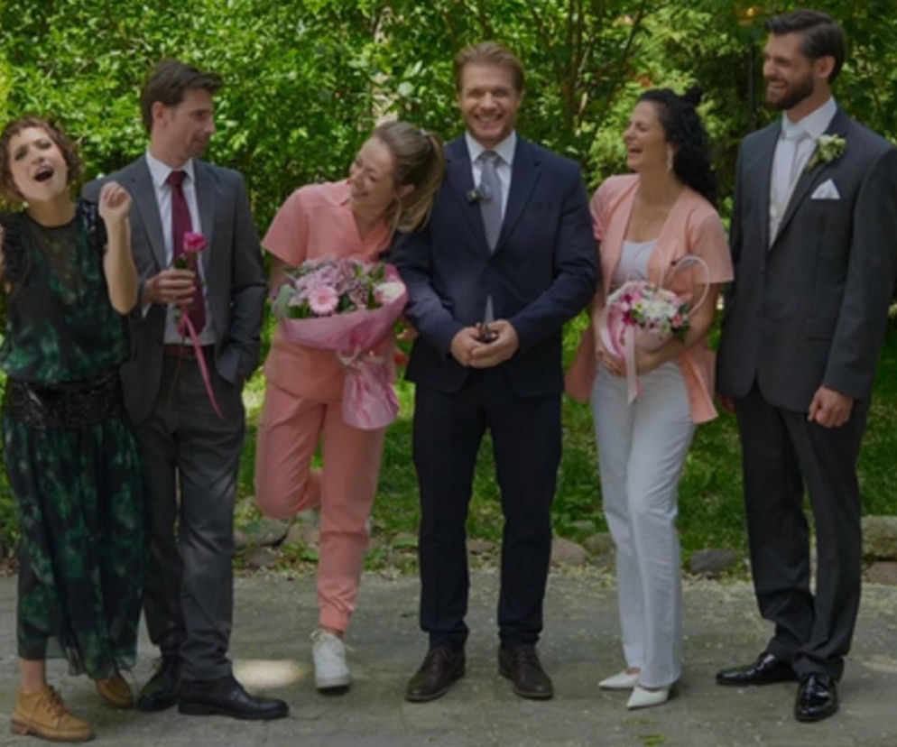 Grupa uśmiechniętych aktorów w odświętnych strojach pozuje w ogrodzie. Tomasz Ciachorowski, Maryla Morydz, Joanna Pach-Żbikowska, Matej Matejka i Aleksandra Hamkało trzymają kwiaty, zapowiadając tajemniczy ślub w nowym sezonie Na dobre i na złe. Więcej szczegółów na temat tej intrygującej fabuły znajdziesz na portalu Super Seriale.