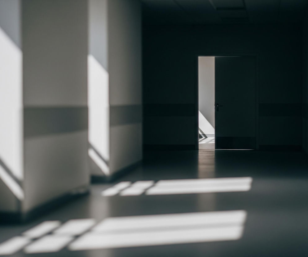 Długi, ciemny korytarz z kremowymi ścianami rozciąga się w głąb obrazu. Z lewej strony światło słoneczne pada przez okno, tworząc jasne, ukośne pasy na ścianie i podłodze. Na końcu korytarza widoczne są otwarte drzwi, przez które wpada jaśniejsze światło, częściowo oświetlając przestrzeń za nimi oraz podłogę. Dolna część ścian po obu stronach korytarza jest ciemniejsza.