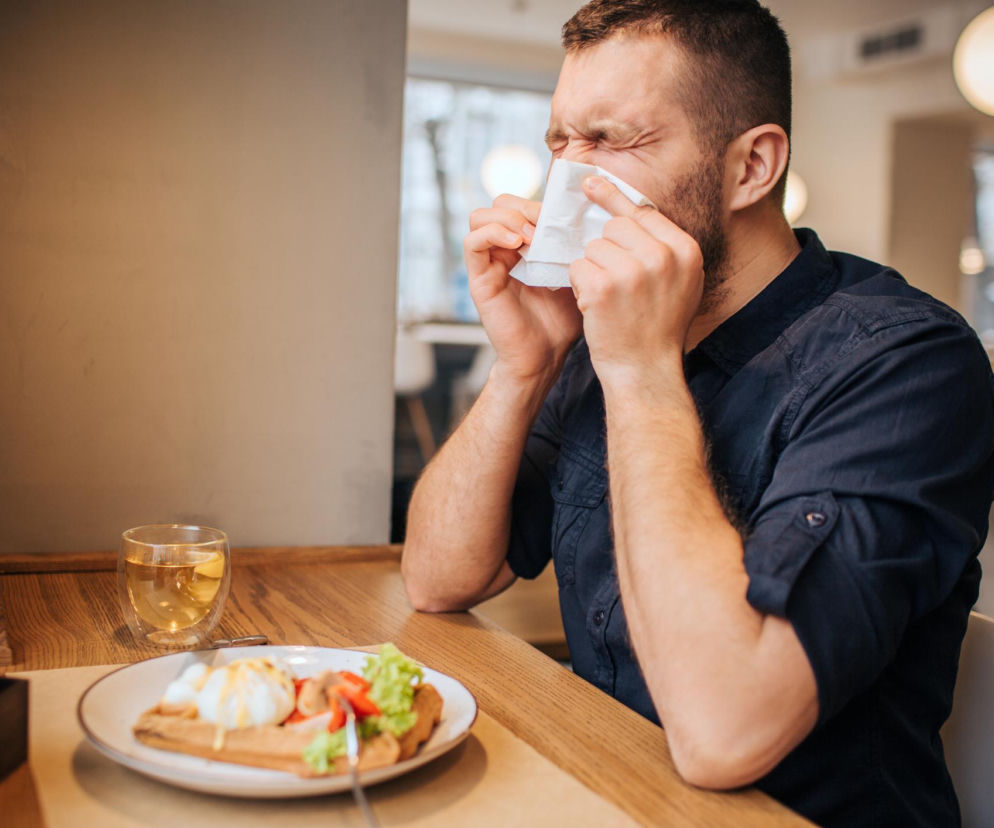 Mężczyzna z katar po jedzeniu, trzymający chusteczkę higieniczną przy nosie, siedzi przy drewnianym stole w restauracji. Na talerzu widoczny posiłek, obok szklanka herbaty. Problem kataru po jedzeniu może dotyczyć nawet kilku milionów Polaków. Przeczytaj więcej na Poradnik Zdrowie.