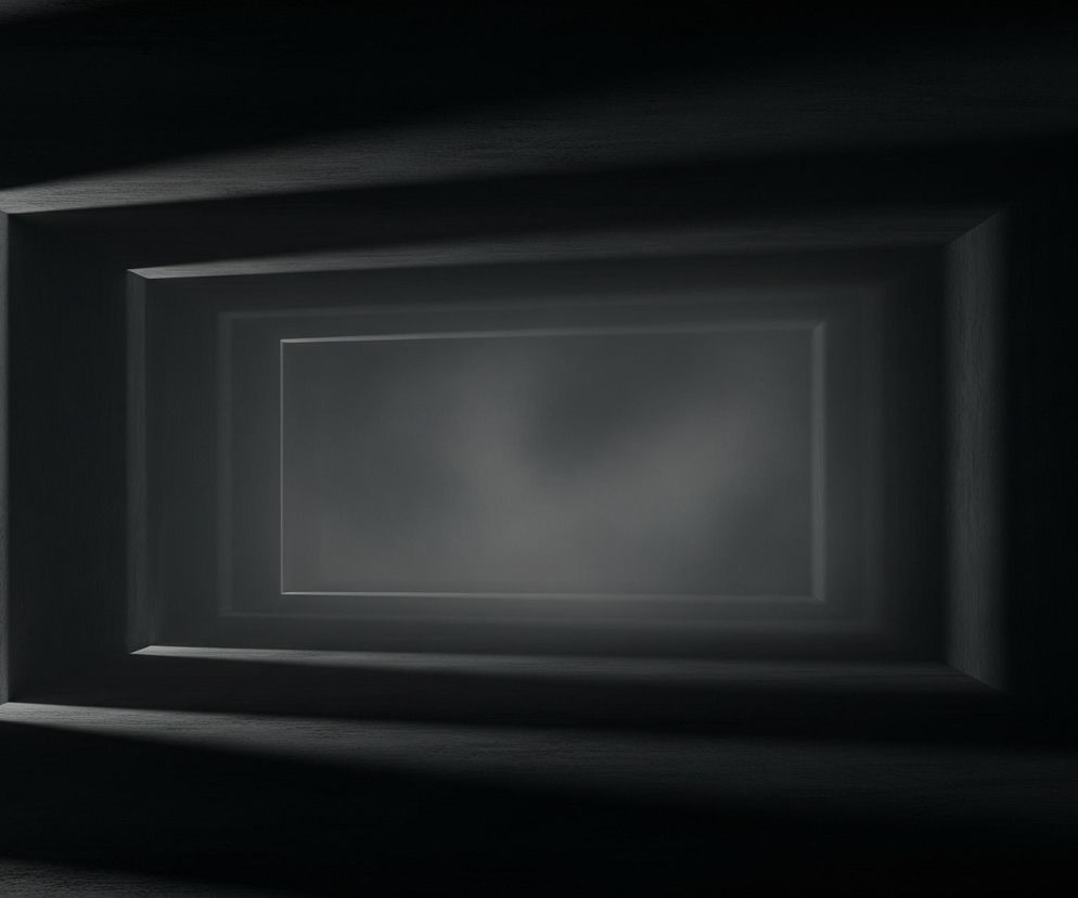 Wizualizacja przedstawia ciemny, pusty tunel lub korytarz o prostokątnym przekroju, w którym ściany, sufit i podłoga mają ciemnoszarą, niemal czarną barwę, z widoczną teksturą przypominającą usłojenie drewna lub chropowatą powierzchnię betonu. Perspektywa prowadzi wzrok w głąb korytarza, gdzie kolejne prostokątne ramy tworzą wrażenie nieskończonej głębi, stając się jaśniejsze i bardziej rozmyte w oddali. Po bokach korytarza i na jego suficie widoczne są subtelne, skośne cienie i smugi światła, akcentujące krawędzie i nadające przestrzeni trójwymiarowości oraz tajemniczego nastroju.