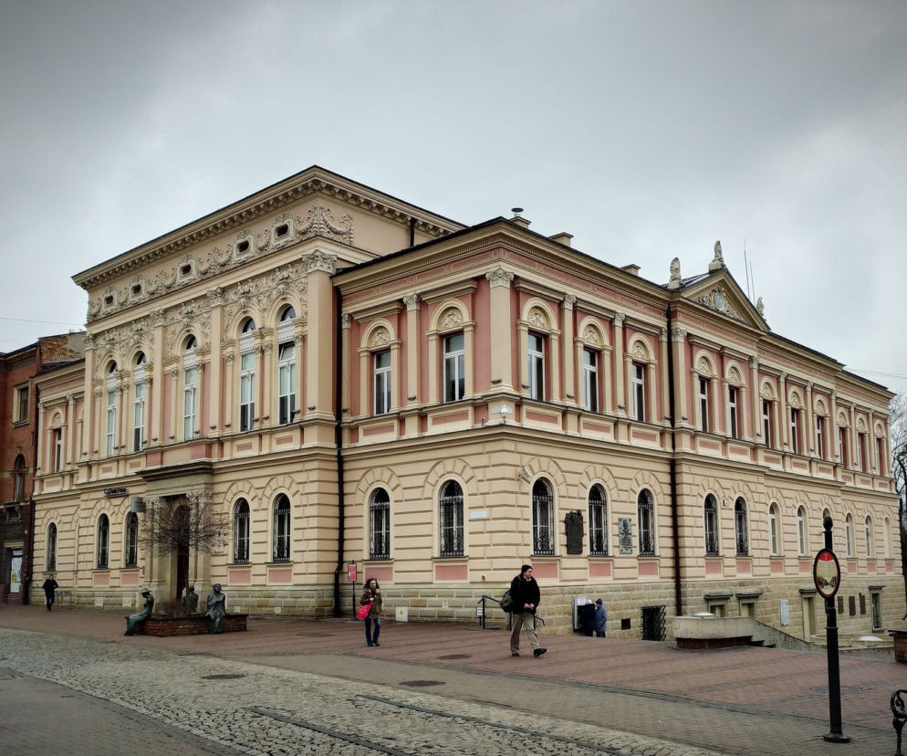 Elegancki, neobarokowy budynek w Tarnowie, z rzeźbionymi fasadami w odcieniach beżu i różu, z charakterystycznymi półokrągłymi oknami na parterze. Przed nim brukowa ulica i kilka osób. Więcej o wyborach w radzie miejskiej przeczytasz na naszym portalu.