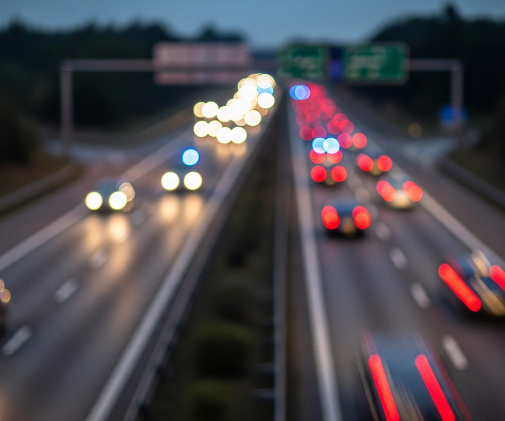 Autostradę w nocy lub o zmierzchu, z rozmyciem typu bokeh, tworzącym efekt świetlistych kręgów. Głównymi elementami są światła samochodów: jasne, żółto-białe kręgi reflektorów i niebieskie światła pojazdu na pierwszym planie po lewej stronie, oraz czerwone kręgi świateł tylnych samochodów w prawej, dalszej części jezdni. Dwupasmowa droga z pasami ruchu oddzielonymi białymi liniami rozciąga się w głąb obrazu, a po obu stronach widać ciemne, rozmyte sylwetki drzew lub zalesionego terenu. W oddali, nad autostradą, widoczne są rozmazane znaki drogowe w kształcie prostokątów z niewyraźnymi, jasnymi napisami.