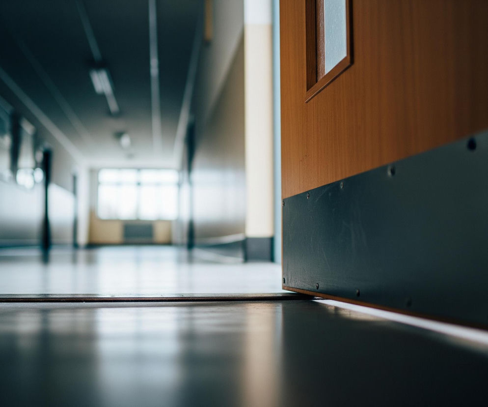 Półotwarte drewniane drzwi z ciemną, metalową listwą u dołu znajdują się po prawej stronie kadru, ukazując fragment długiego korytarza. Korytarz charakteryzuje się ciemnoszarym sufitem z prostokątnymi oprawami świetlnymi oraz jasnymi ścianami, które stopniowo ciemnieją w oddali. Na końcu korytarza widoczne jest duże okno, wpadające przez nie światło tworzy refleksy na polerowanej podłodze.