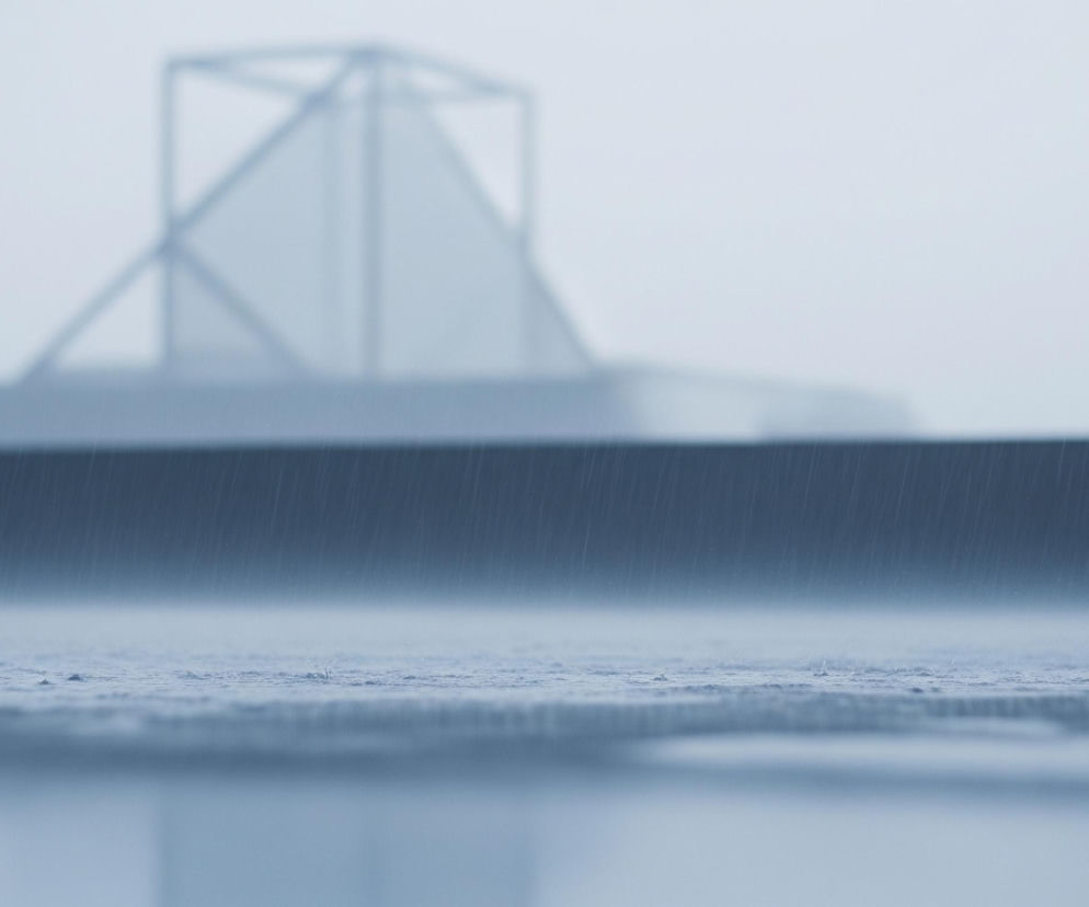 Krople deszczu uderzają o ciemną, poziomą powierzchnię pierwszego planu, tworząc drobne rozbryzgi. Za tą powierzchnią znajduje się kolejna, jaśniejsza, mokra płaszczyzna, również pokryta drobnymi kroplami deszczu. W tle, rozmyte i niewyraźne, rysują się kontury architektonicznych struktur, w tym jasnoszarego, geometrycznego kształtu przypominającego otwartą klatkę lub szkielet budynku na tle jasnego, zamglonego nieba. Całość utrzymana jest w chłodnych odcieniach szarości i błękitu, z dominującą atmosferą deszczowego dnia.