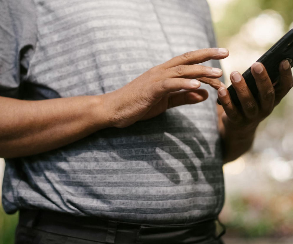 Mężczyzna trzymający czarny smartfon, jego palce dotykają ekranu, a dłonie znajdują się blisko tułowia. Na sobie ma szarą koszulkę w paski, a w tle widać rozmazane tło parku. Artykuł na Poradnik Zdrowie wyjaśnia, jak bezpiecznie używać telefonu po wszczepieniu kardiowertera-defibrylatora.